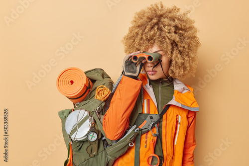 Female explorer looks in binocular explores new places carries big rucksack with necessary things for journey has expedition alone in mountains wears orange jacket. Woman tourist has active lifestyle