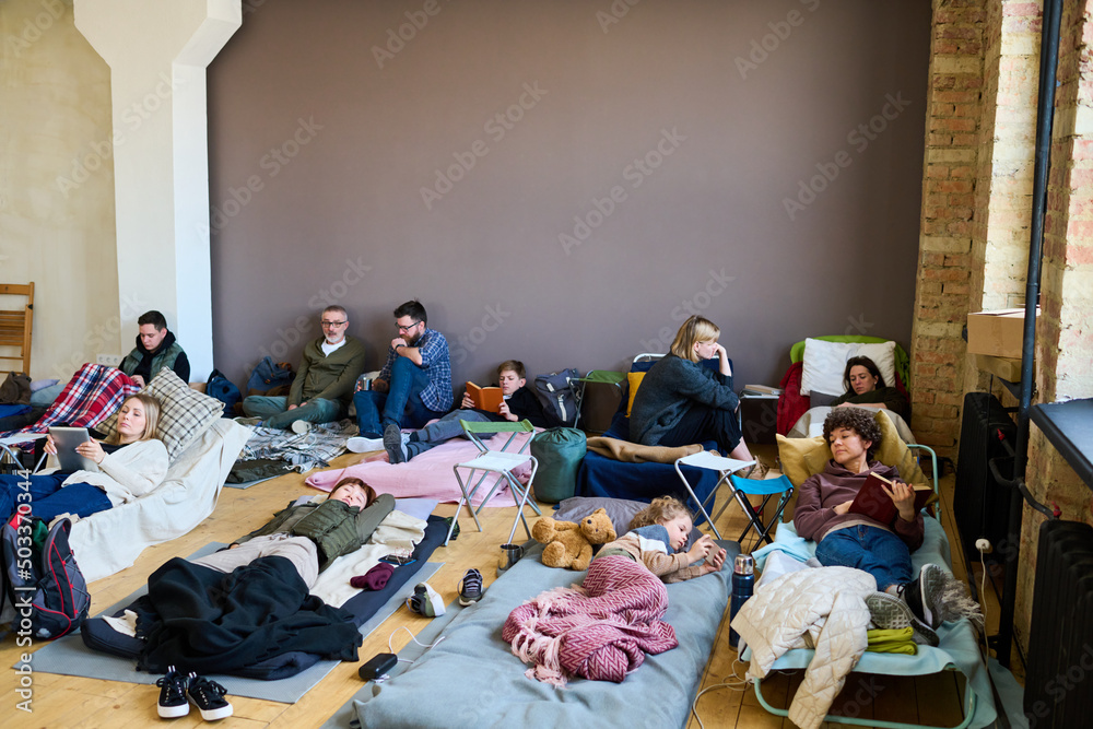 Group of refugees using mobile gadgets, talking and sleeping while having rest on couchettes and mattresses in spacious room - obrazy, fototapety, plakaty 