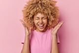 Happy positive emotions concept. Cheerful young woman with cury bushy hair keeps palms raised up exclaims loudly recats to awesome news dressed in casual t shirt isolated over pink background