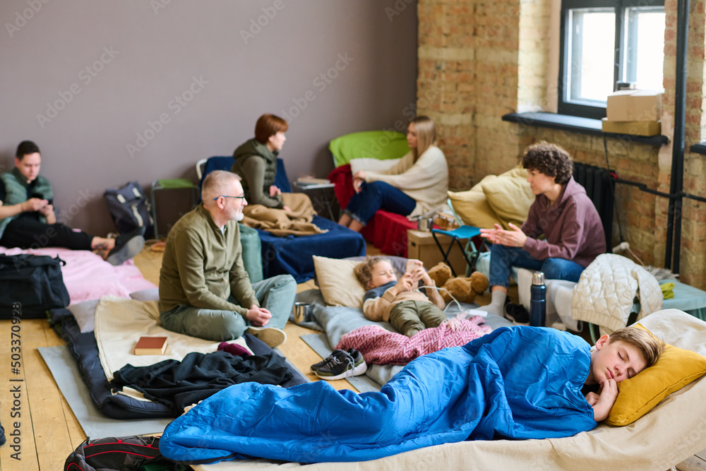 Obraz premium Teenage boy sleeping on couchette against other refugees sitting on their beds and discussing latest news in temporary shelter