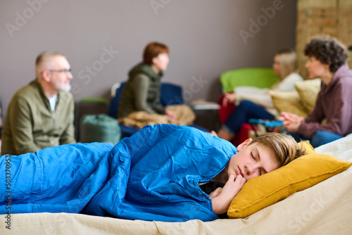 Cute serene boy with his head on pillow sleeping under blanket on couchette against roup of refugees discussing latest news photo