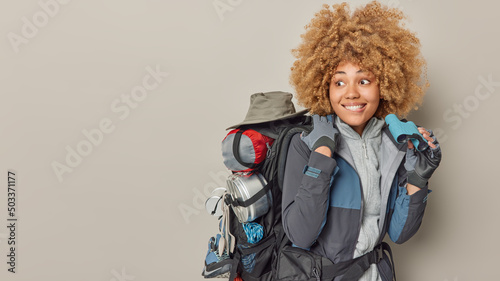 Foto Positive female explorer uses binoculars to look into distance carries backpack has expedition dressed in jacket isolated over grey background copy space for your advertising content