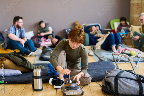 Asian girl in casualwear and eyeglasses cooking food on small electric stove while sitting on mattress in spacious room of refugee camp