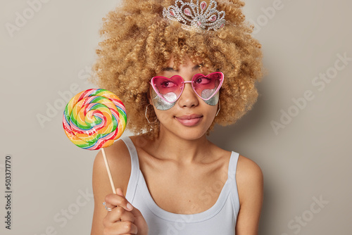 Pensive woman with curly blonde hair wears heart shaped sunglasses applies silver beauty patches under eyes dressed in casual t shirt holds big multicolored lollipop isolated over grey background