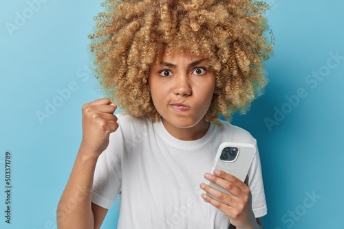 Fotografi Photo of angry outraged curly haired woman purses lips clenches fist looks angrily uses modern mobile phone for sending text messages dressed in casual white t shirt isolated over blue background