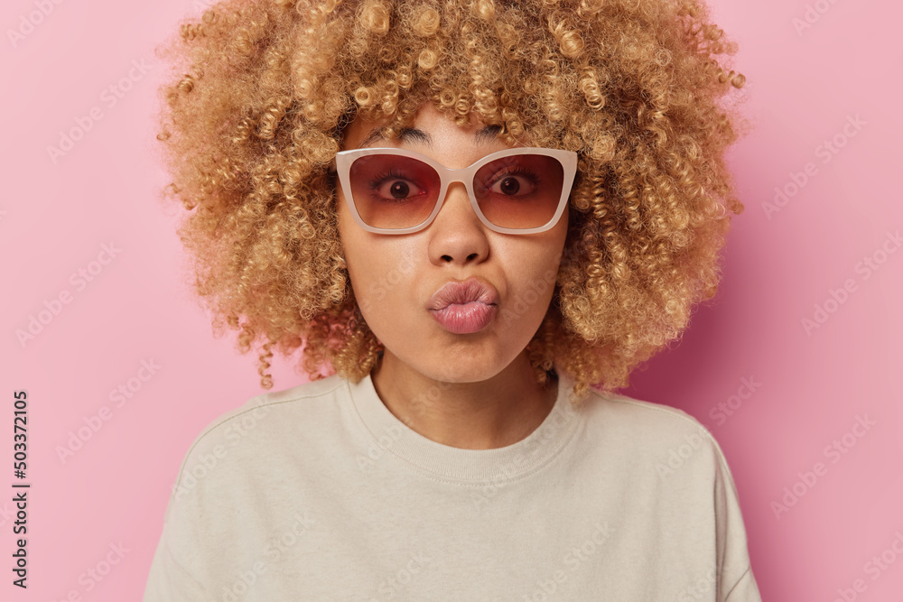 Curly haired woman has romantic mood keeps lips folded wants to kiss you looks through sunglasses dressed in casual t shirt isolated over pink background. Lovely female model blows mwah to lover Curly haired woman has romantic mood keeps lips folded wants to kiss you looks through sunglasses dressed in casual t shirt isolated over pink background. Lovely female model blows mwah to lover