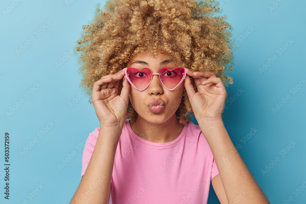Portrait of lovely curly haired woman keeps hands on rim of pink heart sunglasses dressed in casual t shirt isolated over blue background sends air kiss to lover. Human facial expressions concept Portrait of lovely curly haired woman keeps hands on rim of pink heart sunglasses dressed in casual t shirt isolated over blue background sends air kiss to lover. Human facial expressions concept