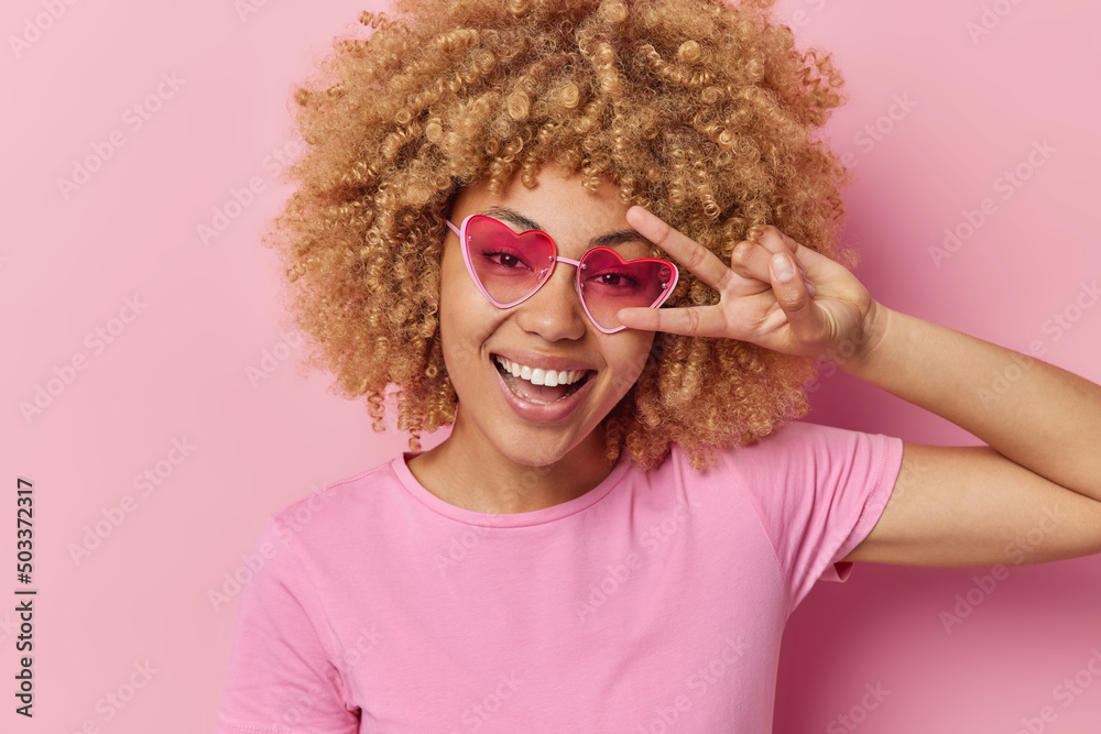 Portrait of happy curly haired woman makes peace gesture over eye smiles toothily has glad expression foolishes around wears heart sunglasses and casual t shirt isolated over pink background.