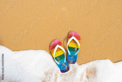 LGBTQ flag flip-flop in a sand and a ocean wave. Top view of a wave in a sandals photo