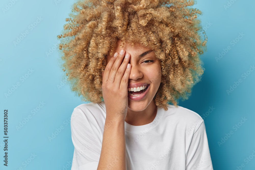 Portrait of carefree young pretty woman with curly hair covers half of face with palm laughs out gladfully dressed in casual white t shirt isolated over blue background. Happy emotions concept Portrait of carefree young pretty woman with curly hair covers half of face with palm laughs out gladfully dressed in casual white t shirt isolated over blue background. Happy emotions concept