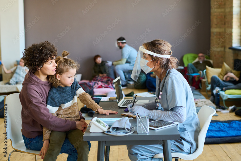Young female refugee with little son on her knees listening to medical advice of doctor in uniform, protective mask and screen - obrazy, fototapety, plakaty 