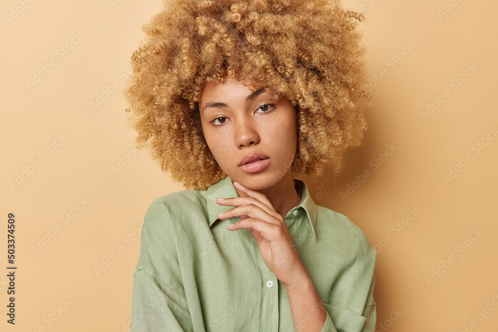 Portrait of serious curly haired woman touches jawline gently looks directly at camera has healthy skin dressed in casual shirt isolated over brown background. Tender female model poses indoors Portrait of serious curly haired woman touches jawline gently looks directly at camera has healthy skin dressed in casual shirt isolated over brown background. Tender female model poses indoors