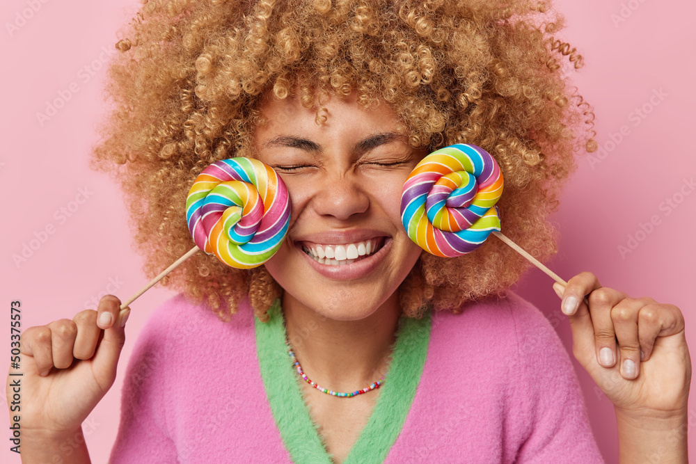Optimistic curly haired young woman keeps two lollipops near face has sweet tooth remembers taste of childhood enjoys eating cheat meal poses against pink background. Sugar harmful for health