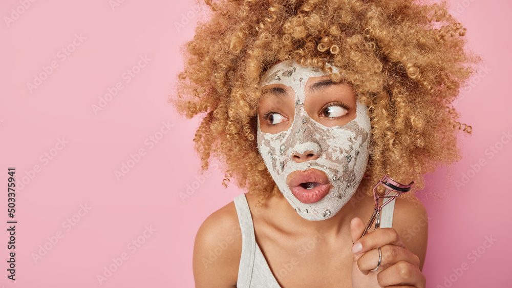 Close up shot of surprised young woman with blonde curly hair applies beauty mask holds eyelashes curler looks amazed away keeps mouth opened stands bare shoulders isolated over pink background Close up shot of surprised young woman with blonde curly hair applies beauty mask holds eyelashes curler looks amazed away keeps mouth opened stands bare shoulders isolated over pink background