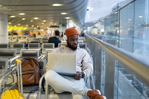 Focused African American man sitting in hall of airport terminal using laptop, wear headphones and listening music. Passenger travelling with backpack, online working on computer and waiting landing. 