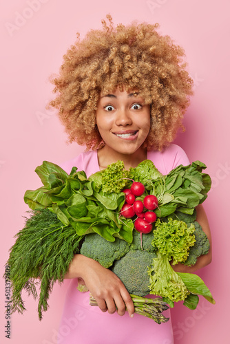 Vertical shot of surprised woman with curly hair bites lips carries fresh green raw vegeatables from greenhouse bites lips poses against pink background came from market. Vegan food ingredients photo