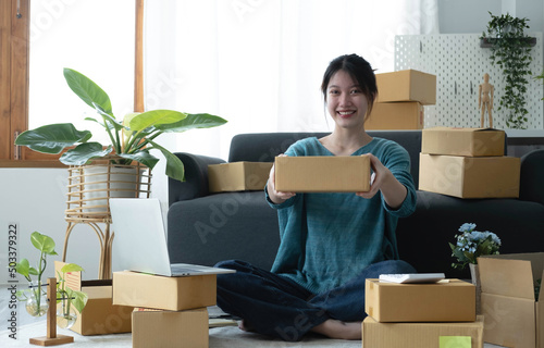 Happy young asian woman startup small business freelance holding parcel box and computer laptop and sitting on floor, Online marketing packing box delivery concept © wichayada