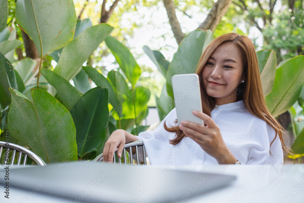 Portrait image of a young asian woman holding and using smart phone with laptop computer on the table in the outdoors Portrait image of a young asian woman holding and using smart phone with laptop computer on the table in the outdoors