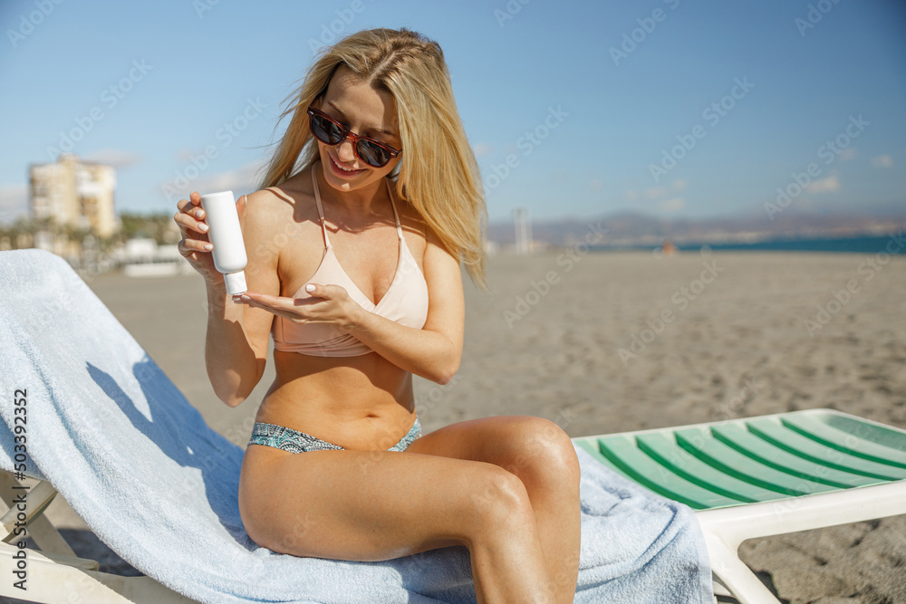 Young lady in swimsuit applying sunscreen cream while sunbathing on beach