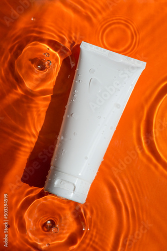 Plastic white tube for cream or lotion on beautiful bursts and glare. Skincare or sunscreen cosmetic on orange background. Beauty concept for face care. Natural sunlight and water spills. Summer mood.