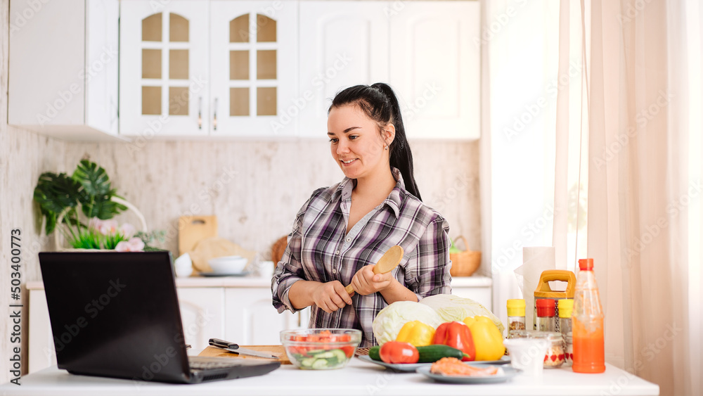 A cheerful young woman cooks in the kitchen with an online audience. The girl stands with a wooden spoon in her hands and looks at her laptop. online cooking.