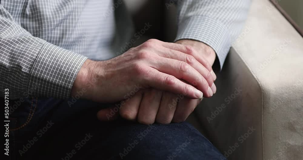 Close up cropped view older man sits on sofa with folded hands feeling upset, recollects past alone indoor, looking lonely. Psychological disorder, elderly age diseases, solitude on retirement concept Close up cropped view older man sits on sofa with folded hands feeling upset, recollects past alone indoor, looking lonely. Psychological disorder, elderly age diseases, solitude on retirement concept