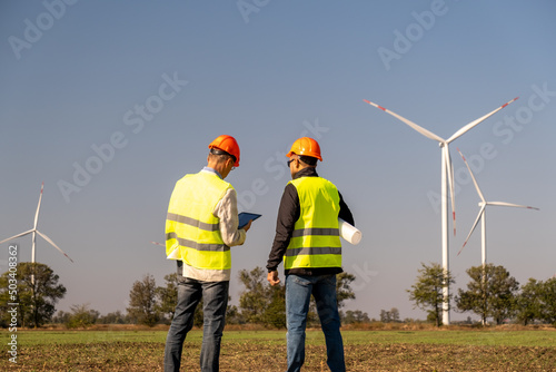 Windmills generate power for national economy in field. Engineers of maintenance control operation of wind farm in countryside via tablet back view