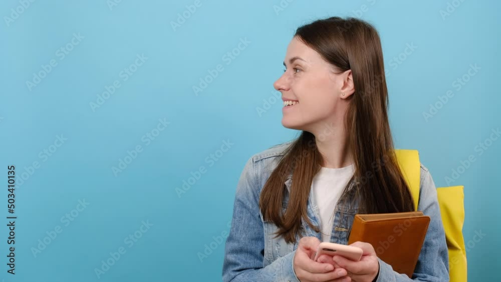 Portrait of smiling girl teen student wear denim jacket and yellow backpack hold books use mobile cell phone look aside on workspace area copy space mock up, isolated on blue studio background wall Portrait of smiling girl teen student wear denim jacket and yellow backpack hold books use mobile cell phone look aside on workspace area copy space mock up, isolated on blue studio background wall