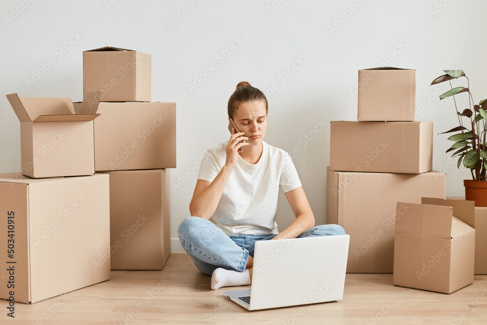 Portrait of Caucasian woman working online on laptop before while moving in a new apartment, talking on mobile phone among cardboard boxes, being upset, posing with pout lips. Portrait of Caucasian woman working online on laptop before while moving in a new apartment, talking on mobile phone among cardboard boxes, being upset, posing with pout lips.