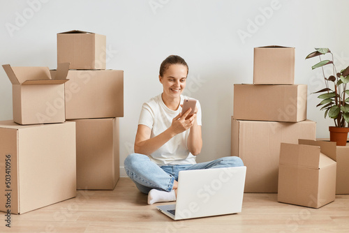 Indoor shot of happy satisfied young adult woman wearing jeans and white t shirt sitting on floor near carton parcels and using phone, working on laptop, expressing positive emotions.