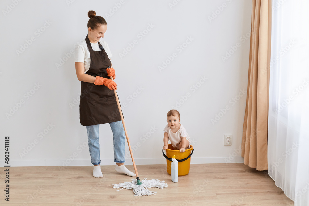 Image of Caucasian young adult housewife cleaning cleaning her house, infection safety, holding mop, cleaning floor, standing near window with her baby in yellow bucket. Image of Caucasian young adult housewife cleaning cleaning her house, infection safety, holding mop, cleaning floor, standing near window with her baby in yellow bucket.
