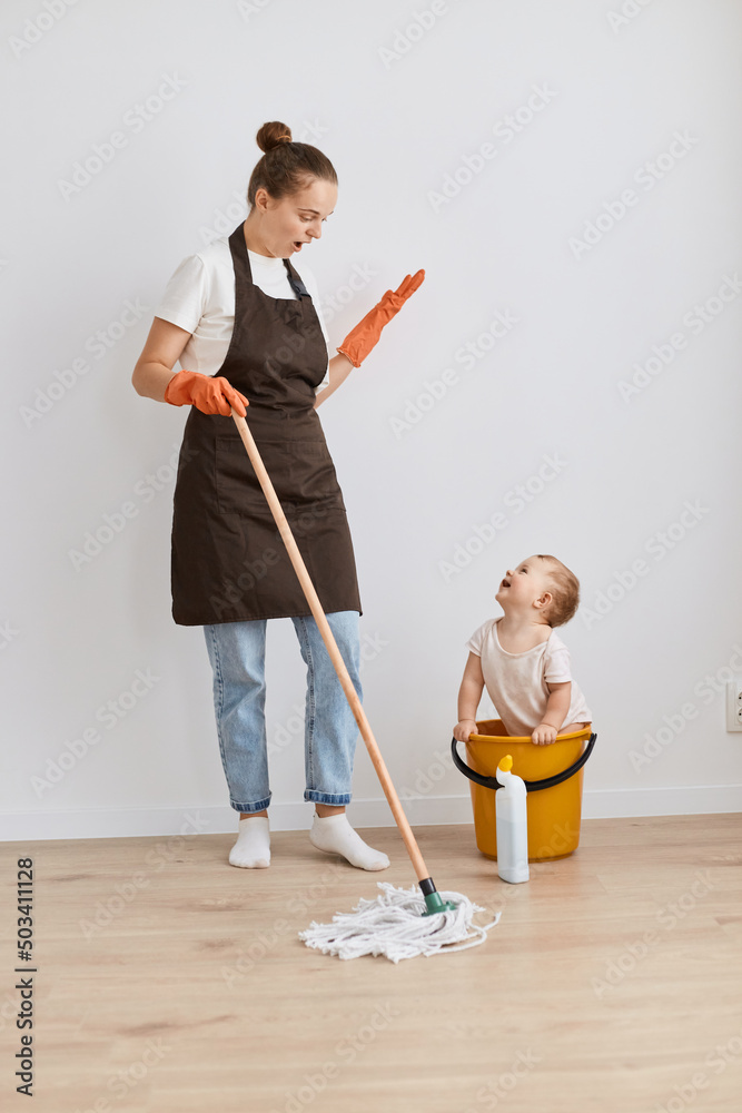 Full length portrait of shocked woman wearing white t shirt and apron, posing with her baby, cleaning her apartment, holding mop, looking at kid in bucket with opened mouth.