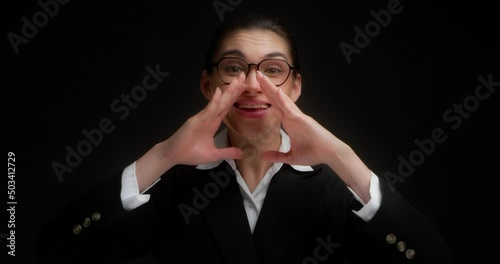 Business woman in glasses and office clothes is looking for you,presses her hands to her mouth,screams loudly.Asian woman wants to share important information.Hey, where are you.On a black background