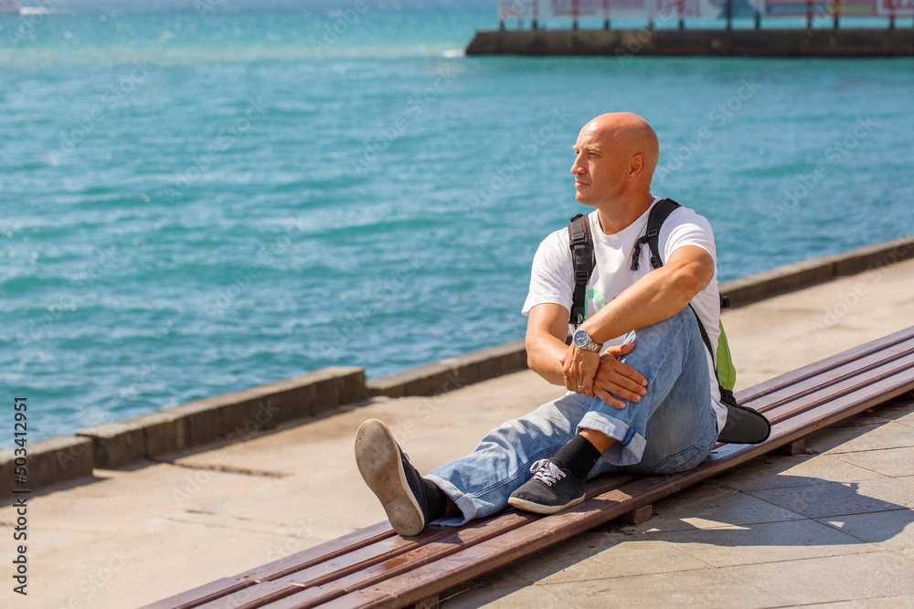Handsome bald head man traveler backpack wearing casual clothes sitting on pavement. A male tourist, relaxing on embankment against the backdrop of the blue sea, enjoys a sunny day. Travel concept