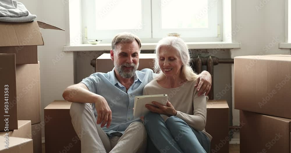 Excited elderly couple sit on floor among boxes with stuff at moving day use pad choose interior design for new flat. Happy aged spouses become homeowners rest from unpacking belongings with tablet pc Excited elderly couple sit on floor among boxes with stuff at moving day use pad choose interior design for new flat. Happy aged spouses become homeowners rest from unpacking belongings with tablet pc