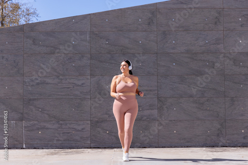 Smiling plus size woman listening music through wireless headphones walking in front of wall