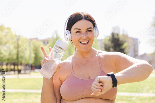Happy plus size woman listening music through wireless headphones holding water bottle showing peace sign in park