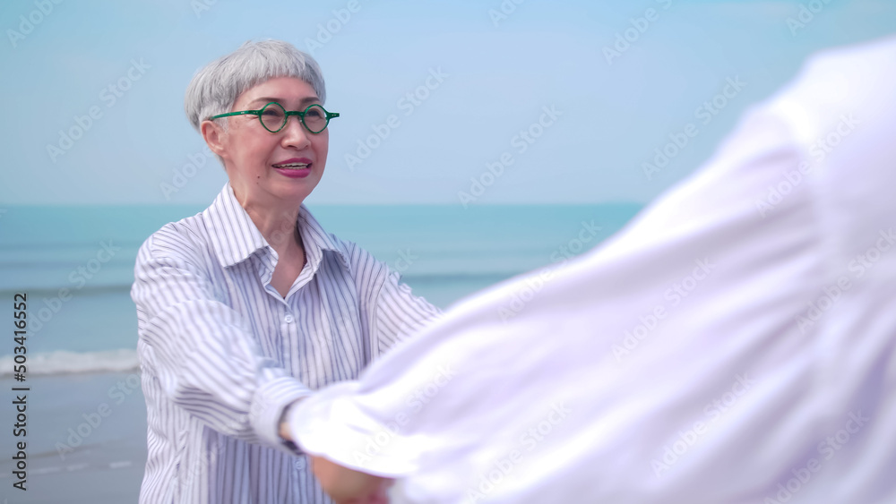Asian retired couple relaxing by the sea in summer. A senior woman dancing with her partner on the beach.