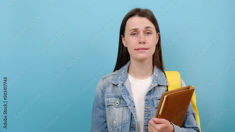 Puzzled pensive girl teen student holding red question marks and books, wears denim jacket and yellow backpack, posing isolated over blue studio background. Education in university college concept