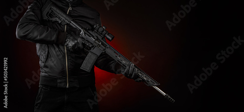 A man in dark clothes holds an automatic carbine in his hands. The face is covered with a mask, hands are in gloves. A figure with a weapon on a dark background with a red light spot.