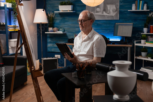 Senior man art creator browsing digital tablet looking for inspiration in online art gallery in home studio with easel. Elderly artist using digital device for improving drawing tehnique in workshop. photo