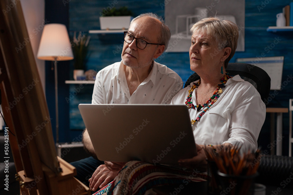 Senior couple sitting in front of easel holding laptop looking at online tutorials to improve drawing technique. Retired man and woman looking for inspiration on social media using portable computer. Senior couple sitting in front of easel holding laptop looking at online tutorials to improve drawing technique. Retired man and woman looking for inspiration on social media using portable computer.