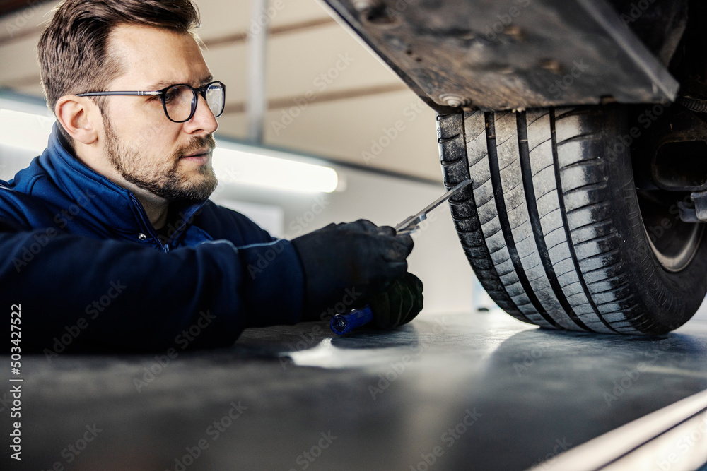 A car mechanic's shop worker checking on car tire in garage. - obrazy, fototapety, plakaty 