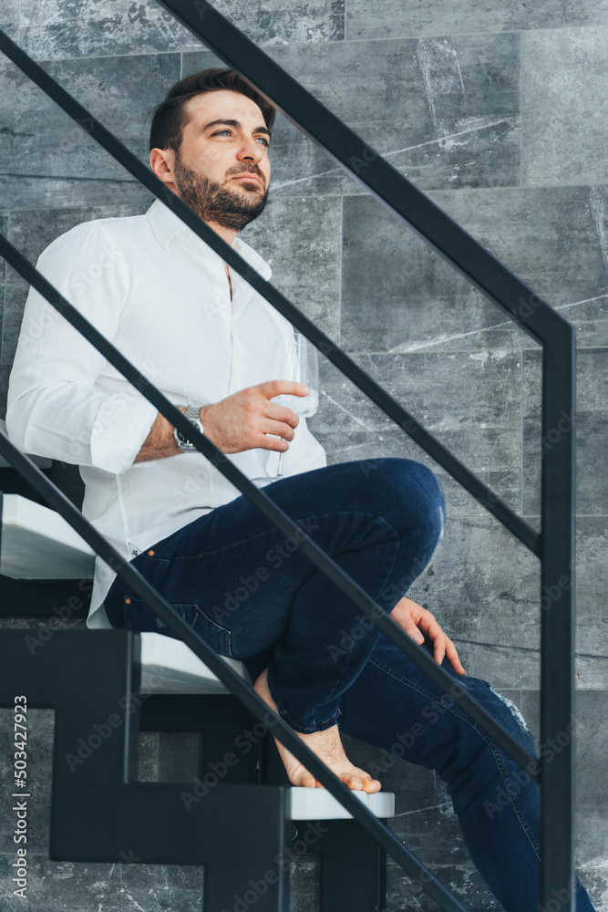 Vertical image of attractive and masculine bearded man sitting on stairs. Holding glass of vine. Promotion, boost career
