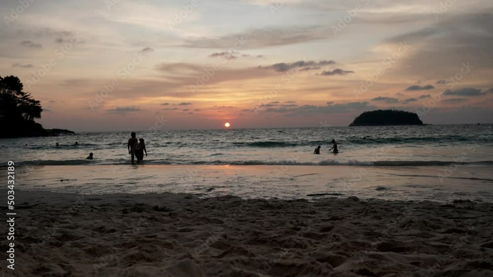 Beautiful timelapse of the beach, sea and clouds. People walk on the sand and admire the departing sun. Many swim in the ocean. An island with trees is visible in the distance. Clouds are floating