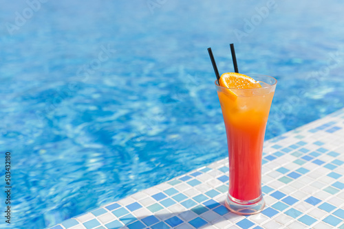 A glass with a red and orange citrus cocktail and two straws stands on the side of the pool