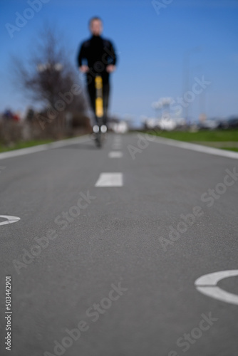 A man on an electric scooter on a bike path. A young man in black clothes on a yellow electric scooter rides on an asphalt bike path with white markings. © Aleksandr