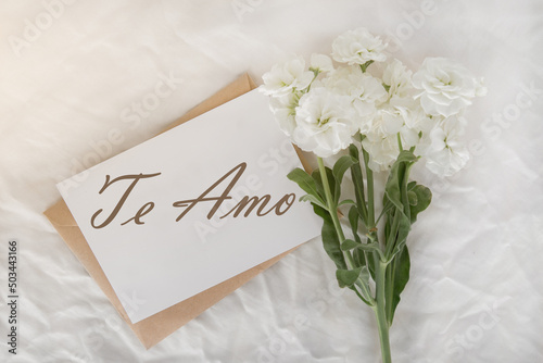 a white small bouquet on a white sheet, a craft envelope and a card with the inscription I love you in Spanish - te amo