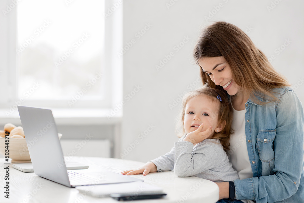 Cute mother hugs baby works from home, she sits in the kitchen and using laptop for remote work, her cute little girl sits nearby on a chair and looks at the computer Cute mother hugs baby works from home, she sits in the kitchen and using laptop for remote work, her cute little girl sits nearby on a chair and looks at the computer