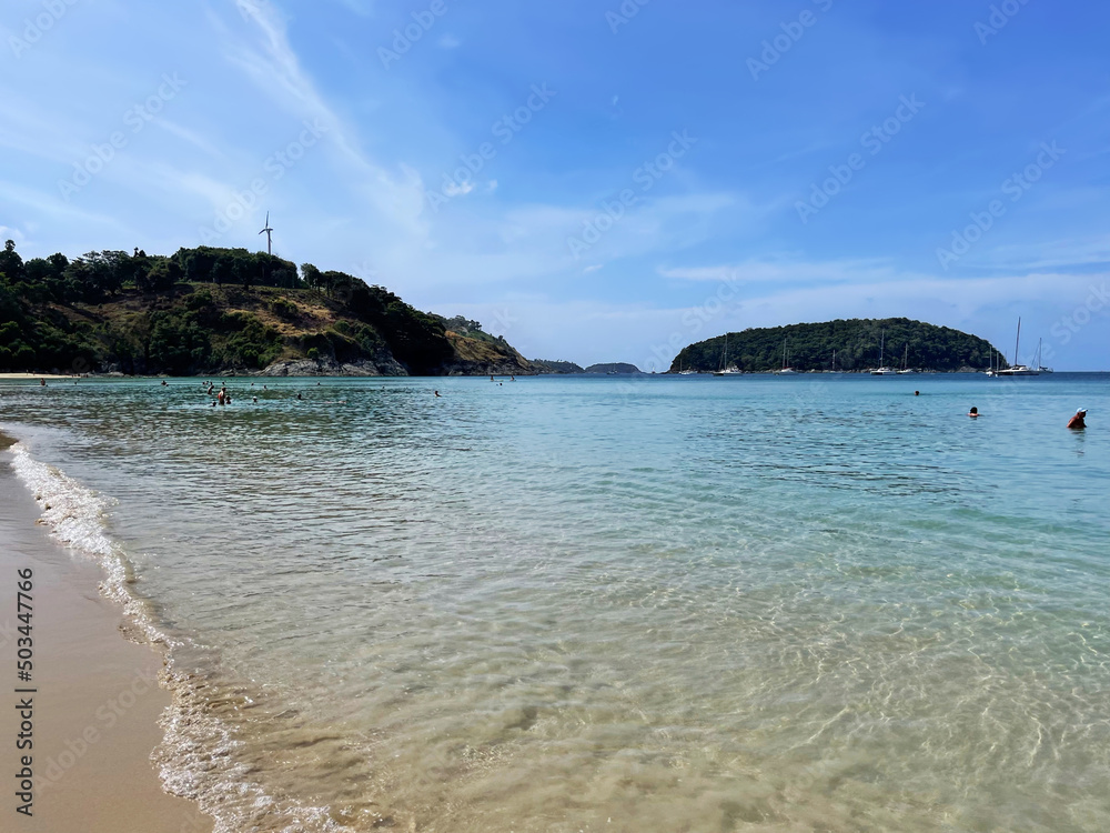 Tropical beach. Transparent water. People swim in calm clear sea. Islands and sailboats in the distance. Wind turbine on top of a mountain. Blue sky and clouds. Landscape. Seascape. Vacationers. Tour 